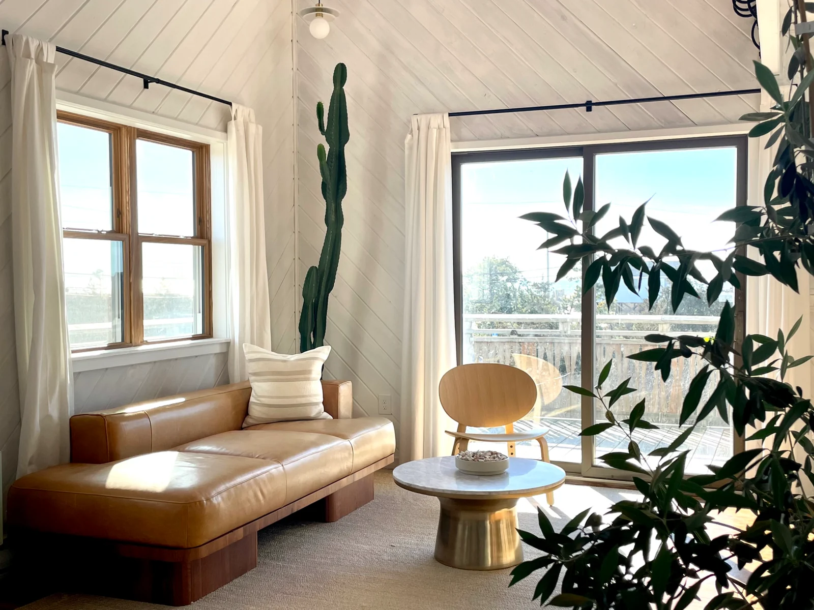 Sunlight across a caramel leather sofa, a wooden lounge chair, and a stone-topped brass table at Surf House